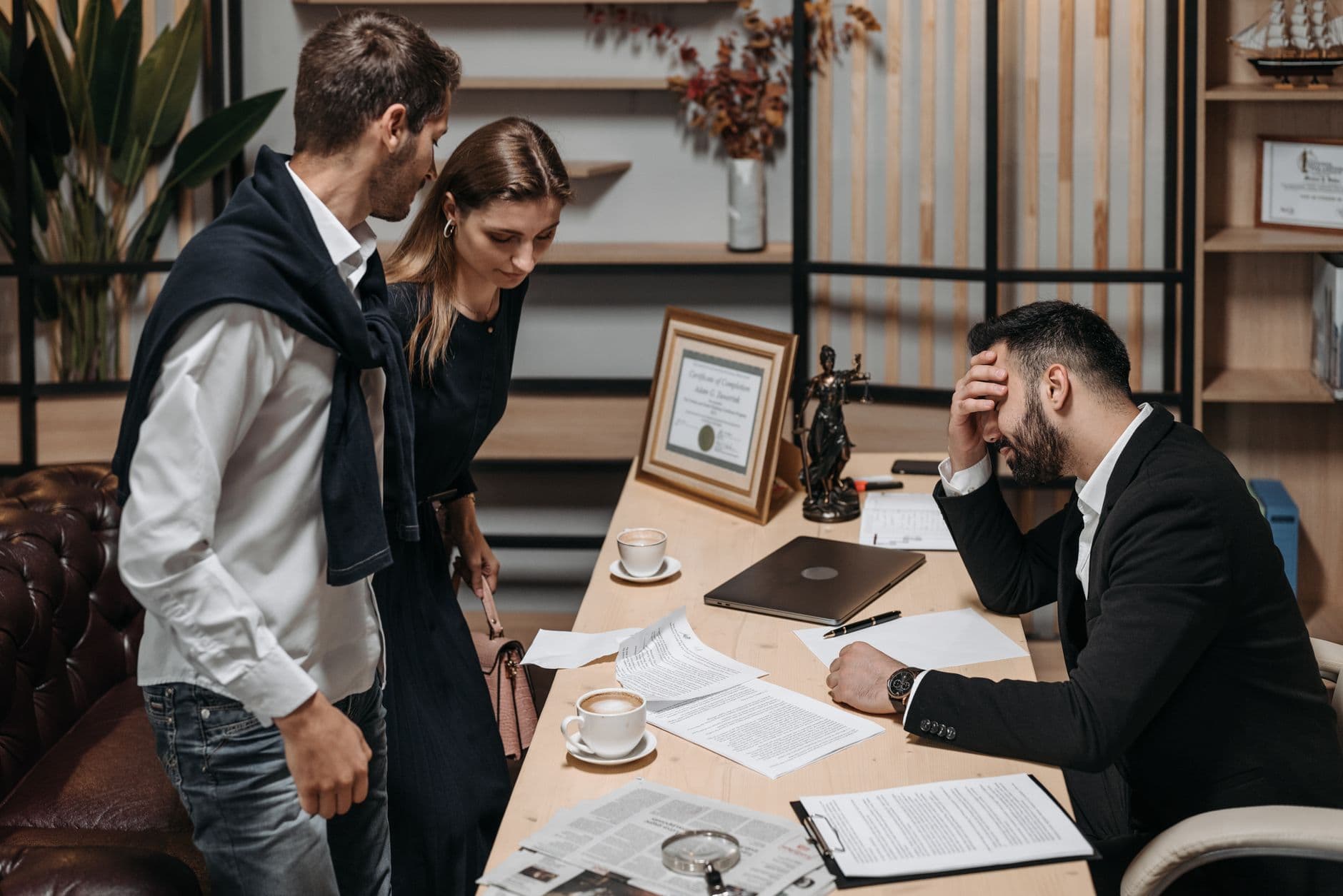 Two colleagues discuss reports with a stressed professional holding his head at an office desk.