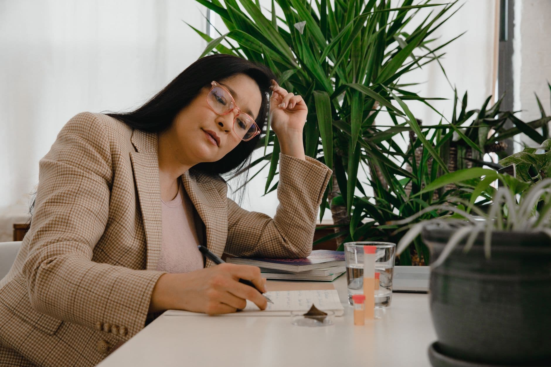 Asian businesswoman with glasses writing in office surrounded by plants.