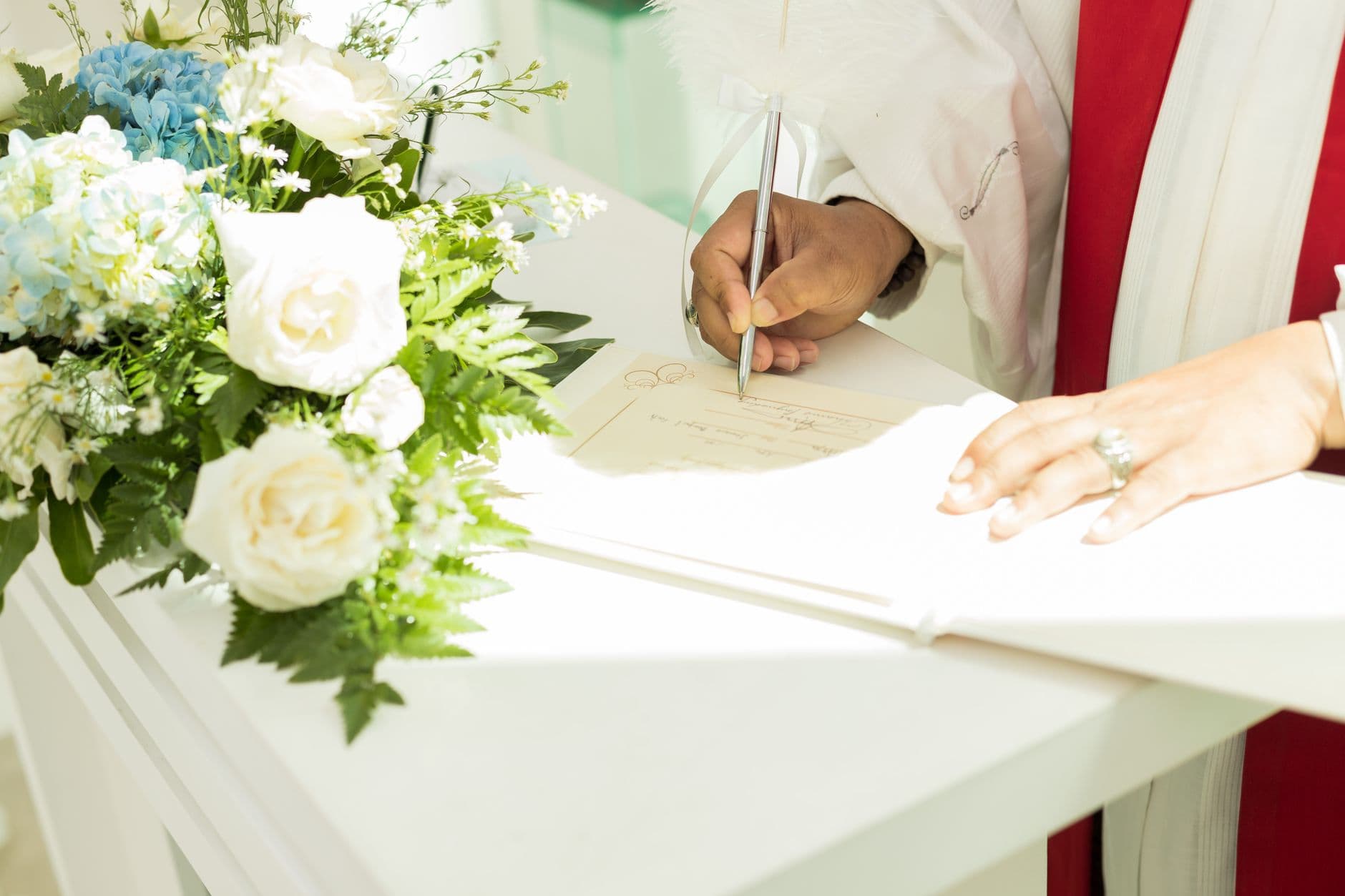 A priest signs an important document next to a bouquet of white roses, creating a serene scene.
