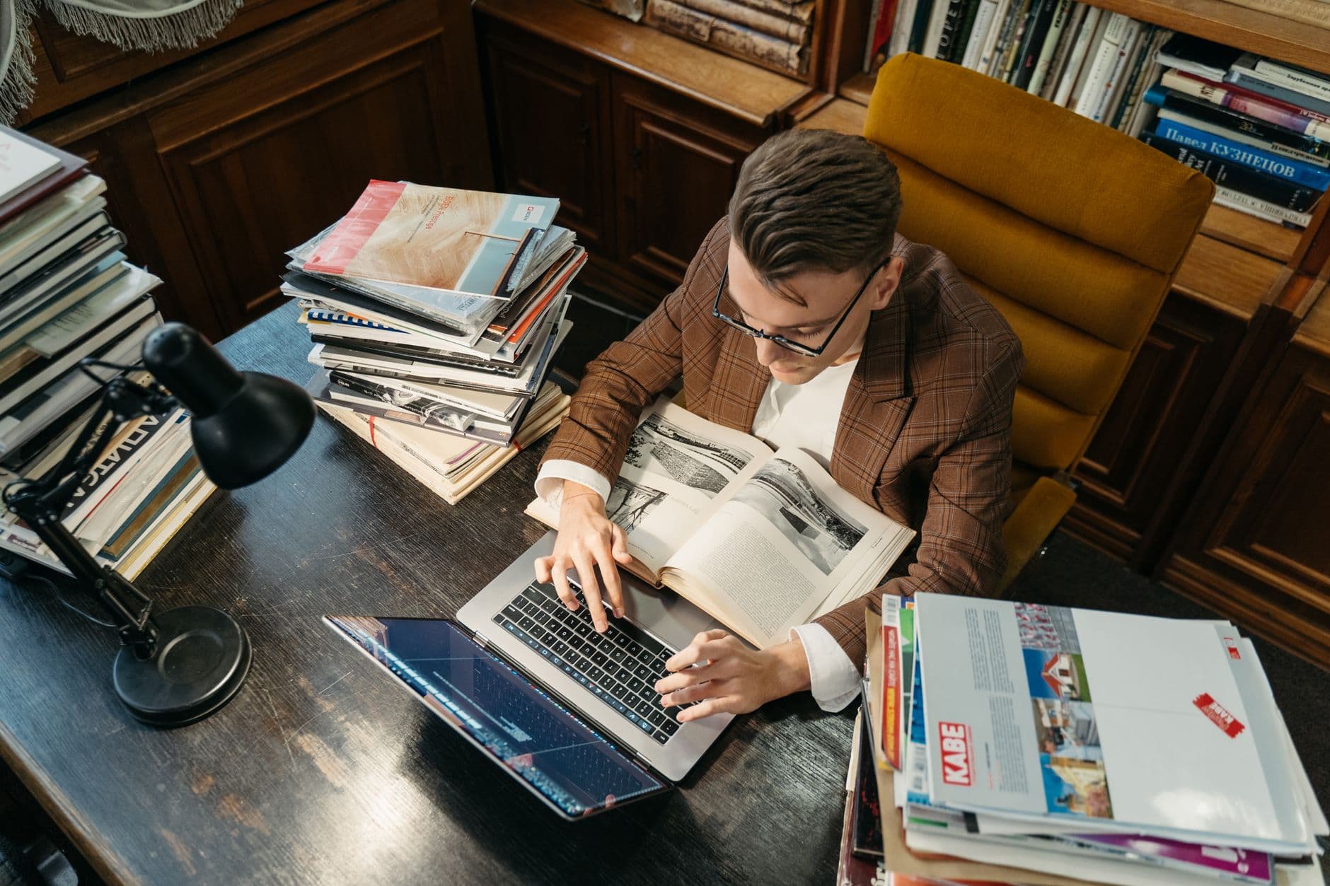 A young man studies in a cozy library setting, using a laptop and books at a wooden desk.